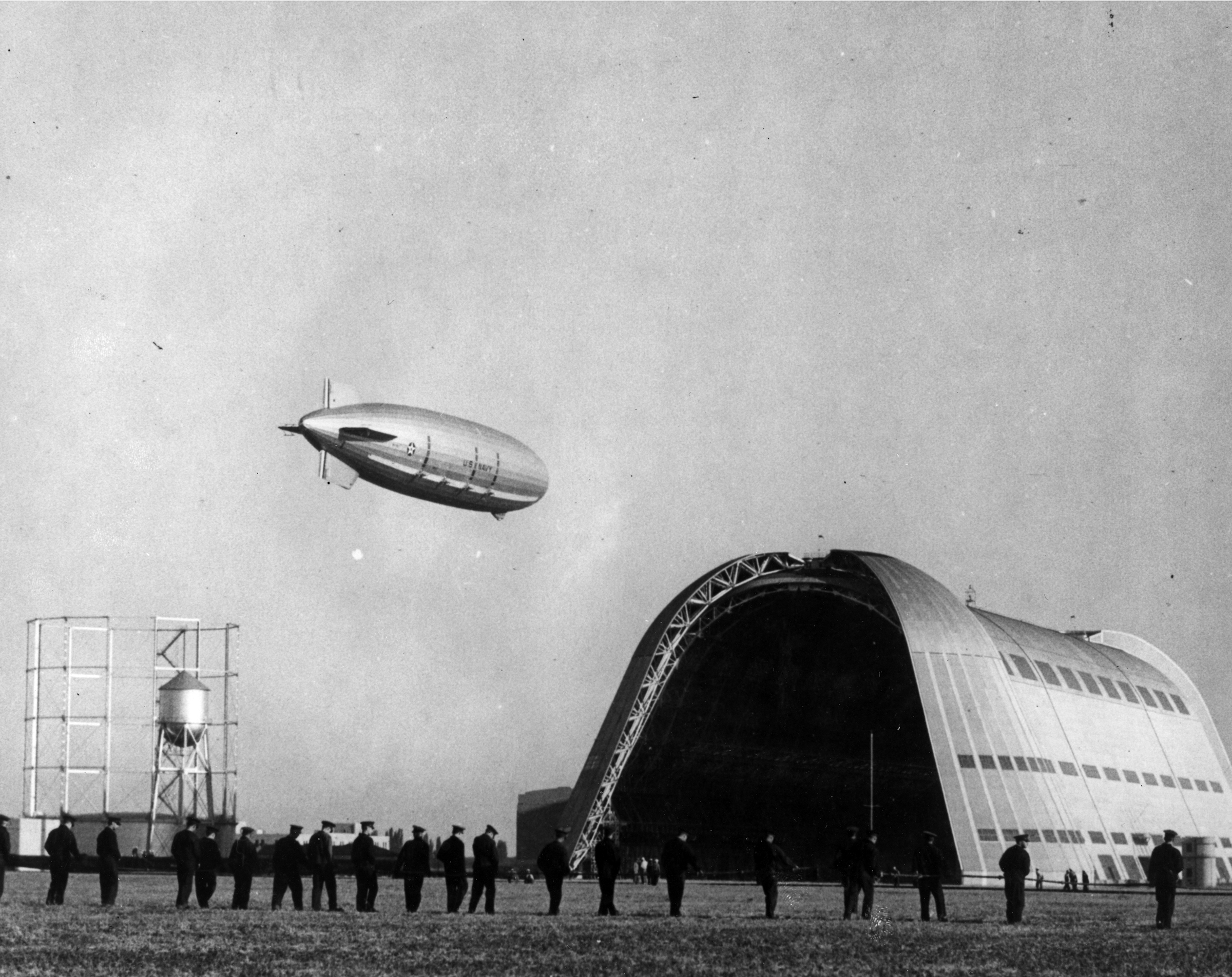 The USS Macon dirigible soars above the huge open doors of Hangar 1, a circular gasholder tower,  water tower, and a line of 21 Navy sailors.