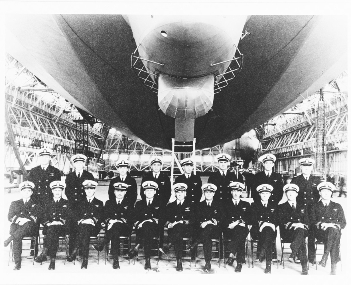 19 Navy officers wearing dark formal uniforms and white hats pose in 2 rows under the USS Macon dirigible moored inside Hangar 1.