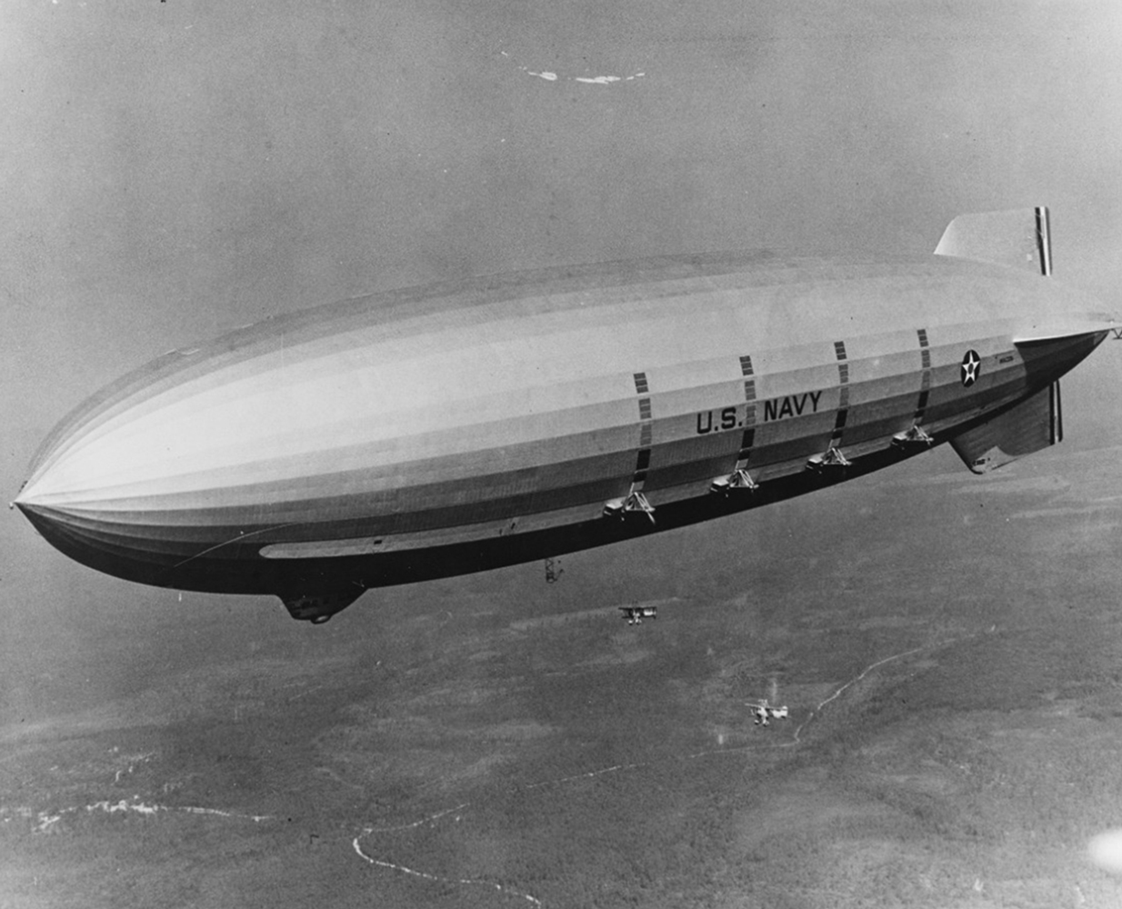 The USS Macon dirigible soars high above an open landscape. Two small planes that were deployed from the belly of the Macon, fly below the airship.