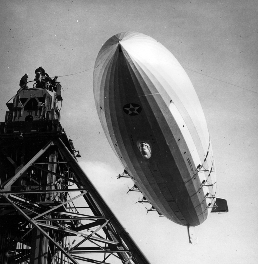 The USS Macon dirigible, seen from below, descending toward a tall mooring mast. Men standing on top of the tower guide a cable attached to the Macon.