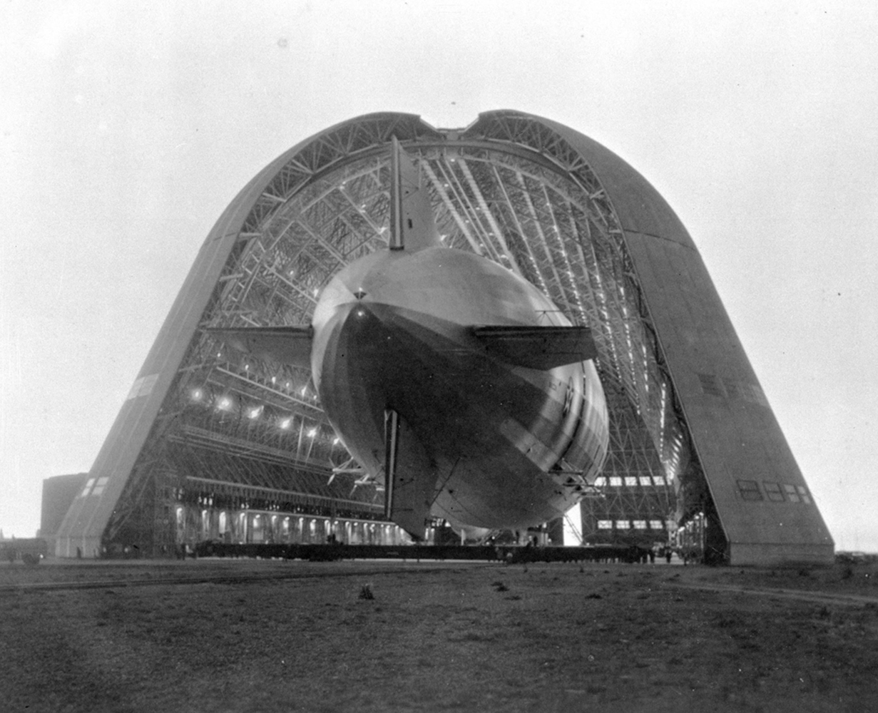 The USS Macon dirigible entering Hangar 1 through its enormous open doors. The inside of the huge hangar is dramatically illuminated.