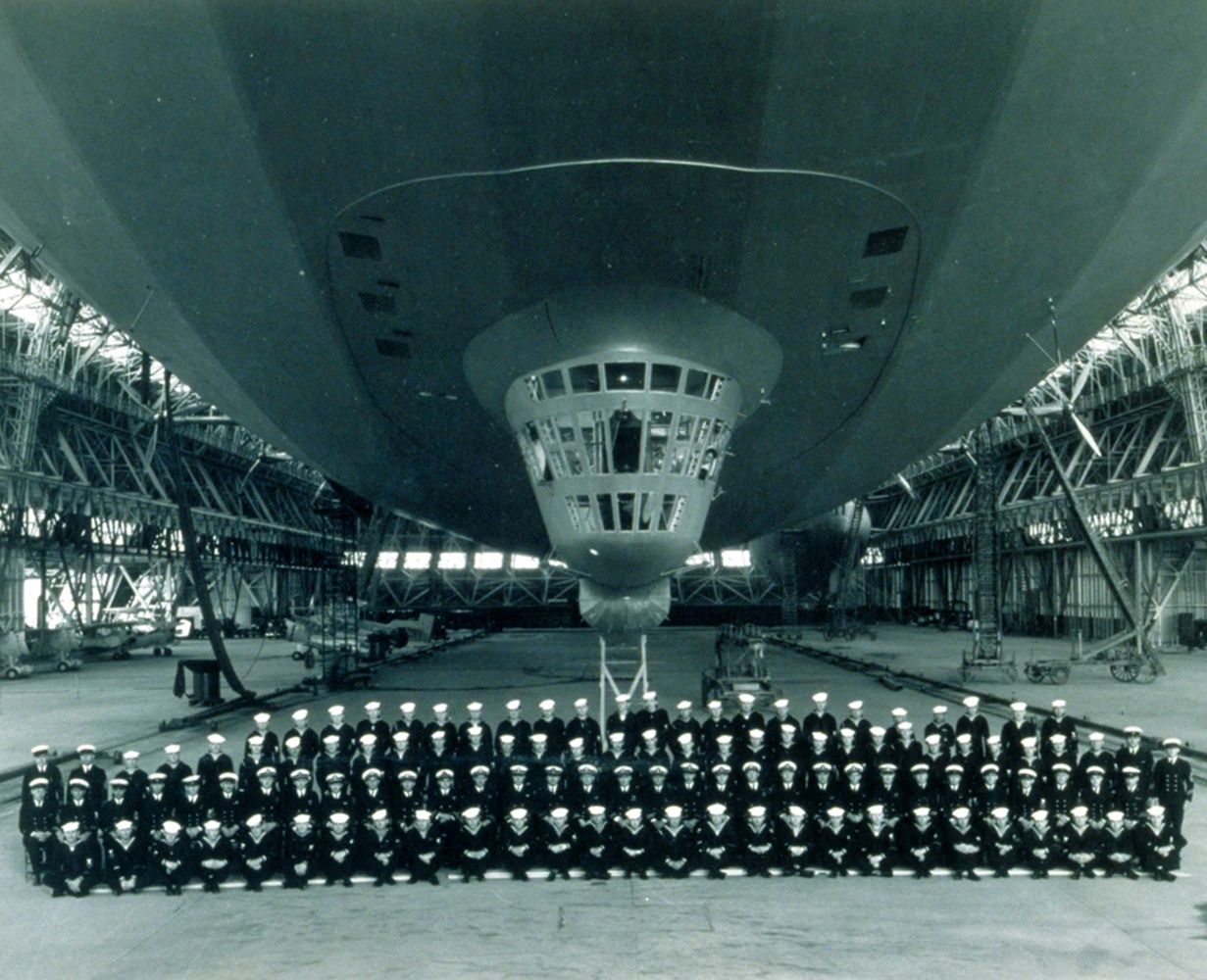 115 Navy officers and crew members pose in 4 rows under the USS Macon dirigible moored inside Hangar 1.