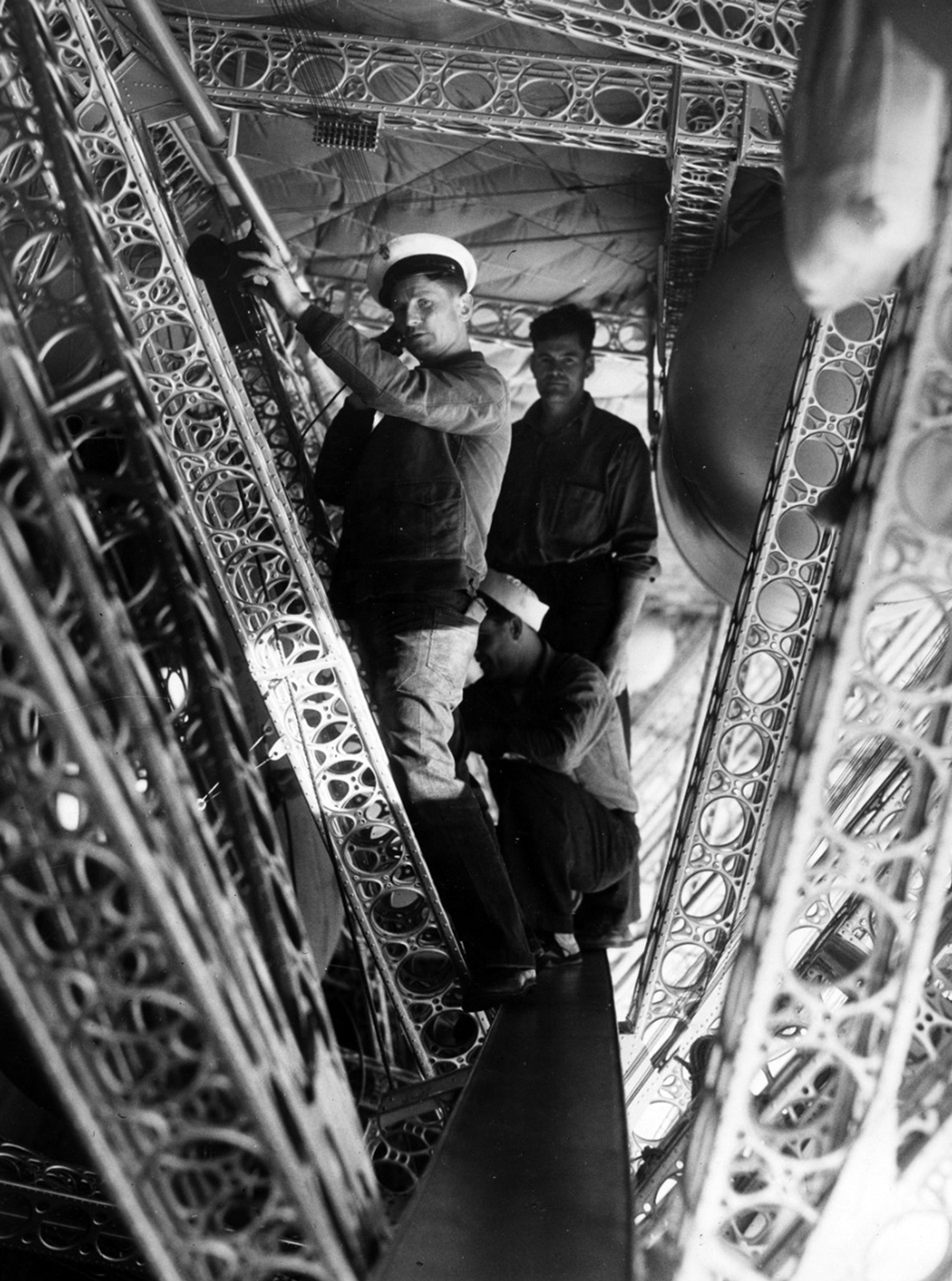 Three crew members of the USS Macon, stand on the narrow catwalk between the metal structural ribs of the huge rigid airship.