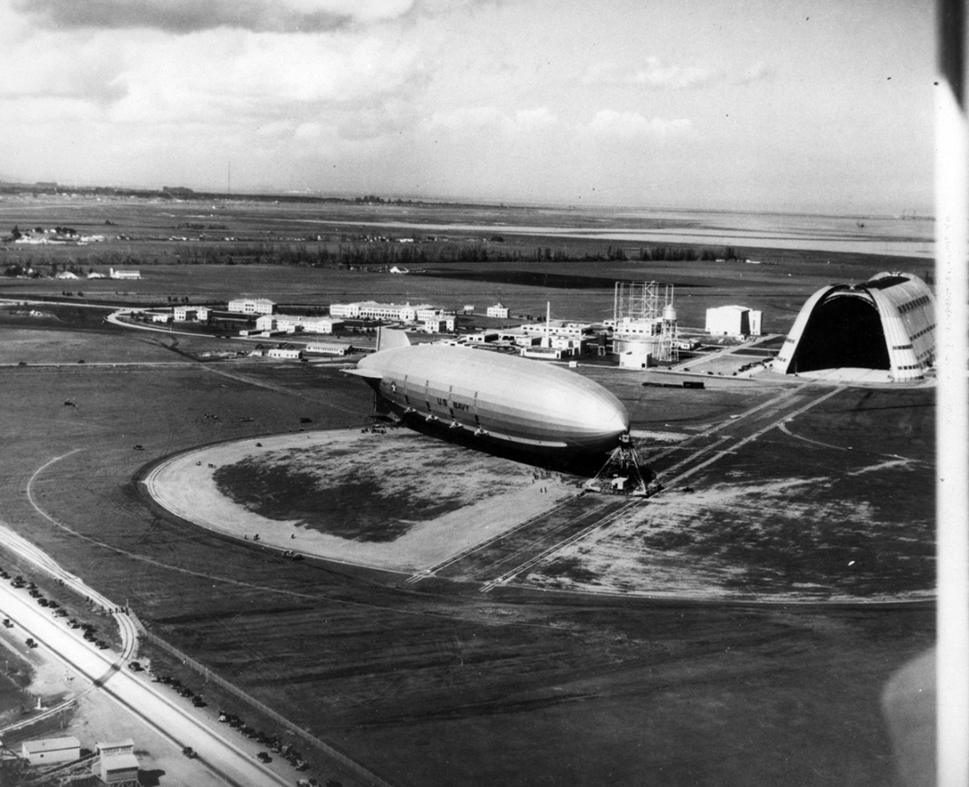 A birds-eye view of the USS Macon dirigible attached to a mooring mast on the airfield near the huge open doors of Hangar 1.