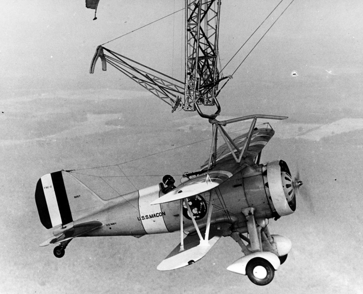 A single propeller biplane, with a pilot in the cockpit, hangs high in the air from the trapeze hook attached to the bottom of the USS Macon dirigible.