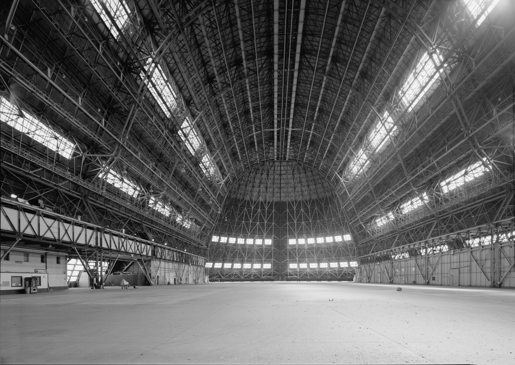 The vast, empty interior space of Hangar 1 and its exposed steel structural system, two rows of long windows, and tall doors at the end of the building.
