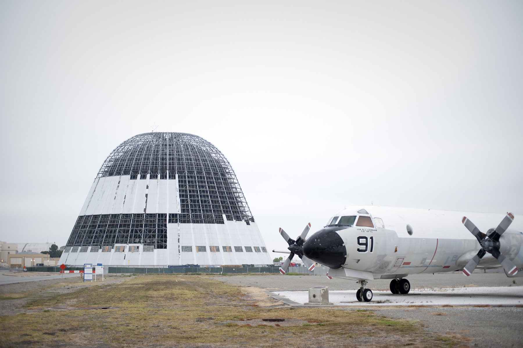The south end of Hangar 1 with most of its metal panels removed and its steel structure exposed. A parked, white, P-3 Orion airplane is on the right. 