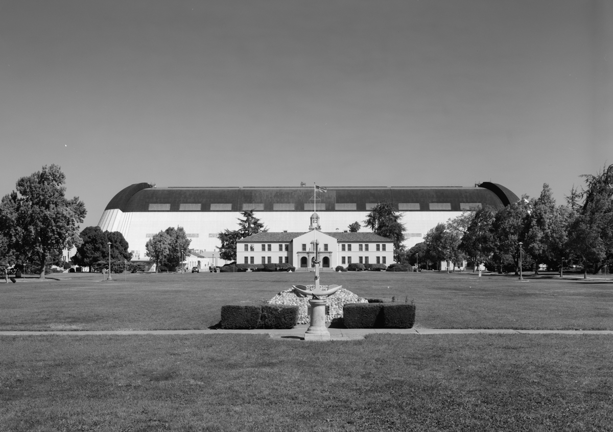An open lawn with a view of the long façade of Hangar 1 behind a building with a tile roof and bell tower. An anchor is displayed in the foreground.