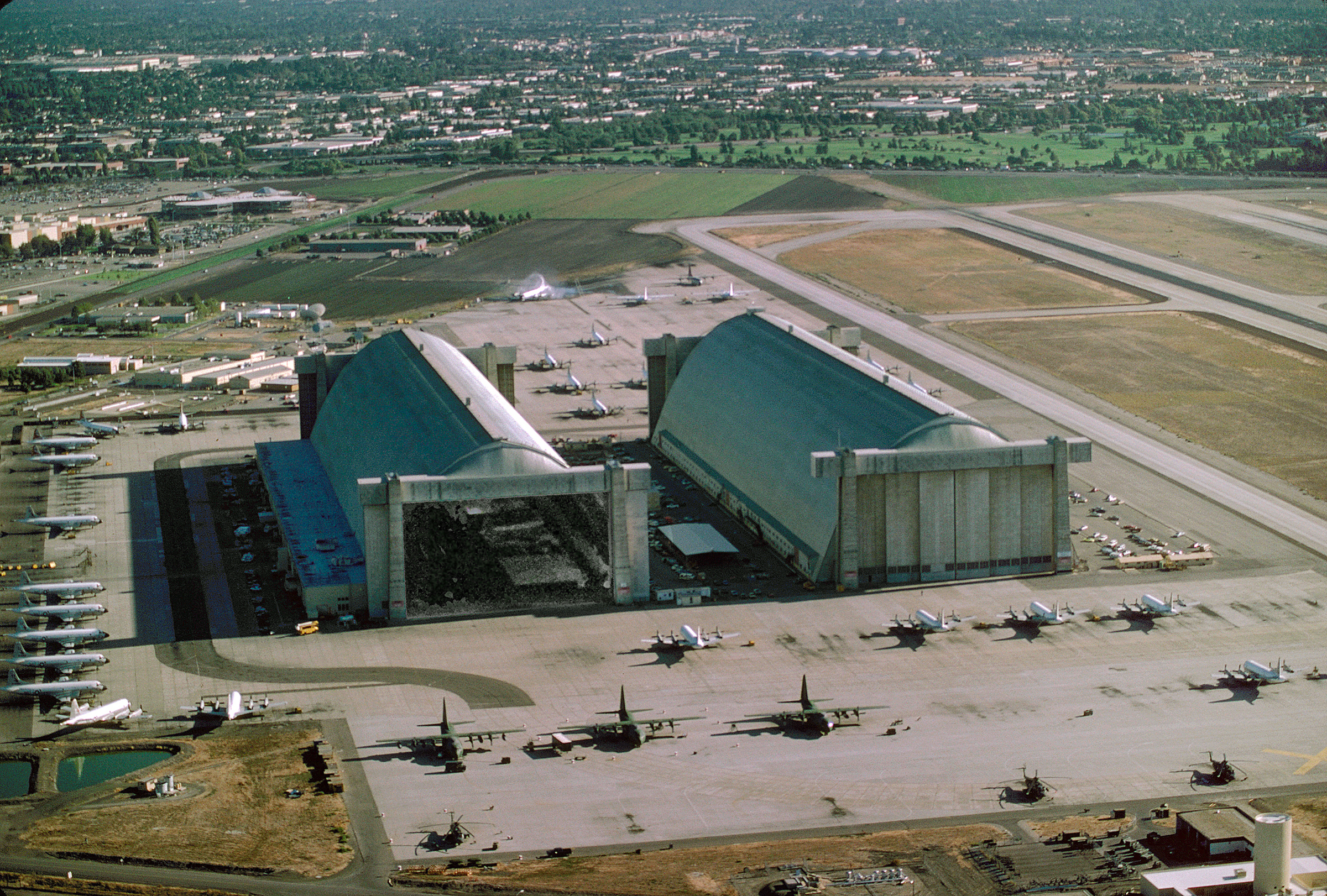 A birds-eye view of the north end of Hangars 3 and 2. The doors of Hangar 3 are open. 3 cargo planes, 3 helicopters, and 28 P-3 planes are parked outside.