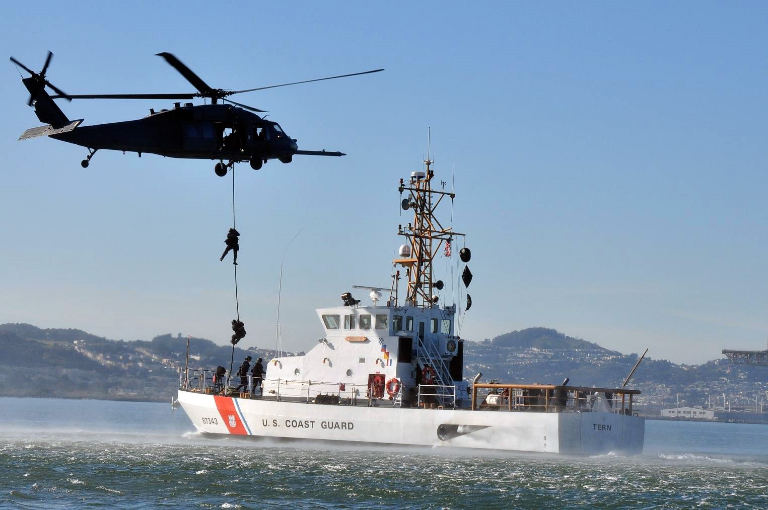 A rescue helicopter hovers above a white ship with U. S. Coast Guard written on its side. 2 people repel down a cable from the helicopter to the ship.