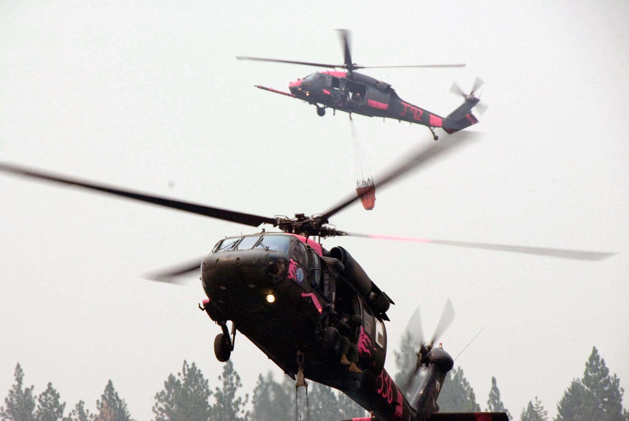 Two rescue helicopters hover above a line of pine trees. A water bucket attached with long cables is seen hanging from the bottom of one helicopter.
