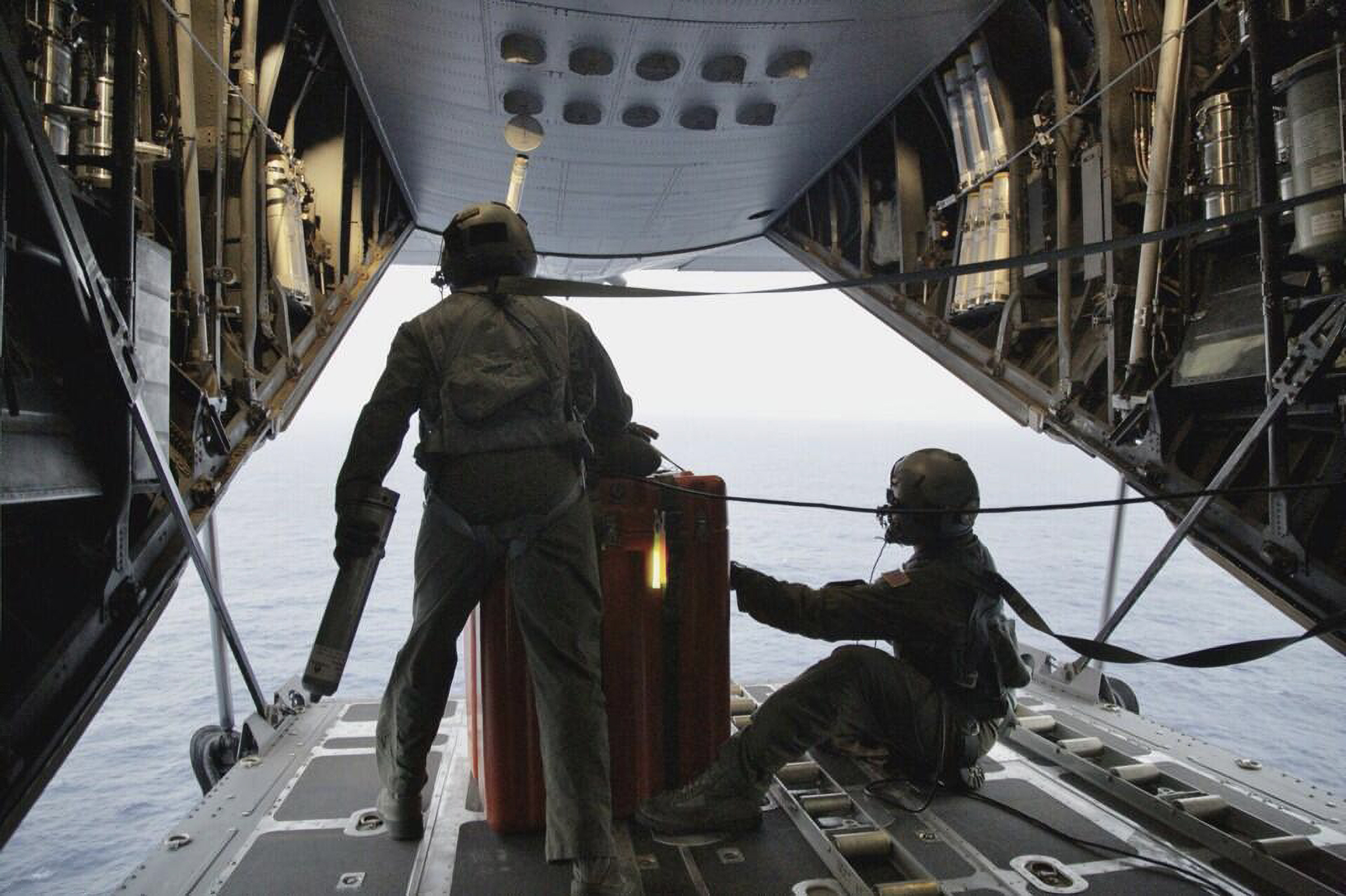 Two pararescuemen, with their backs to the camera, look out of the large, open, back cargo door of a transport plane flying near the surface of the sea.