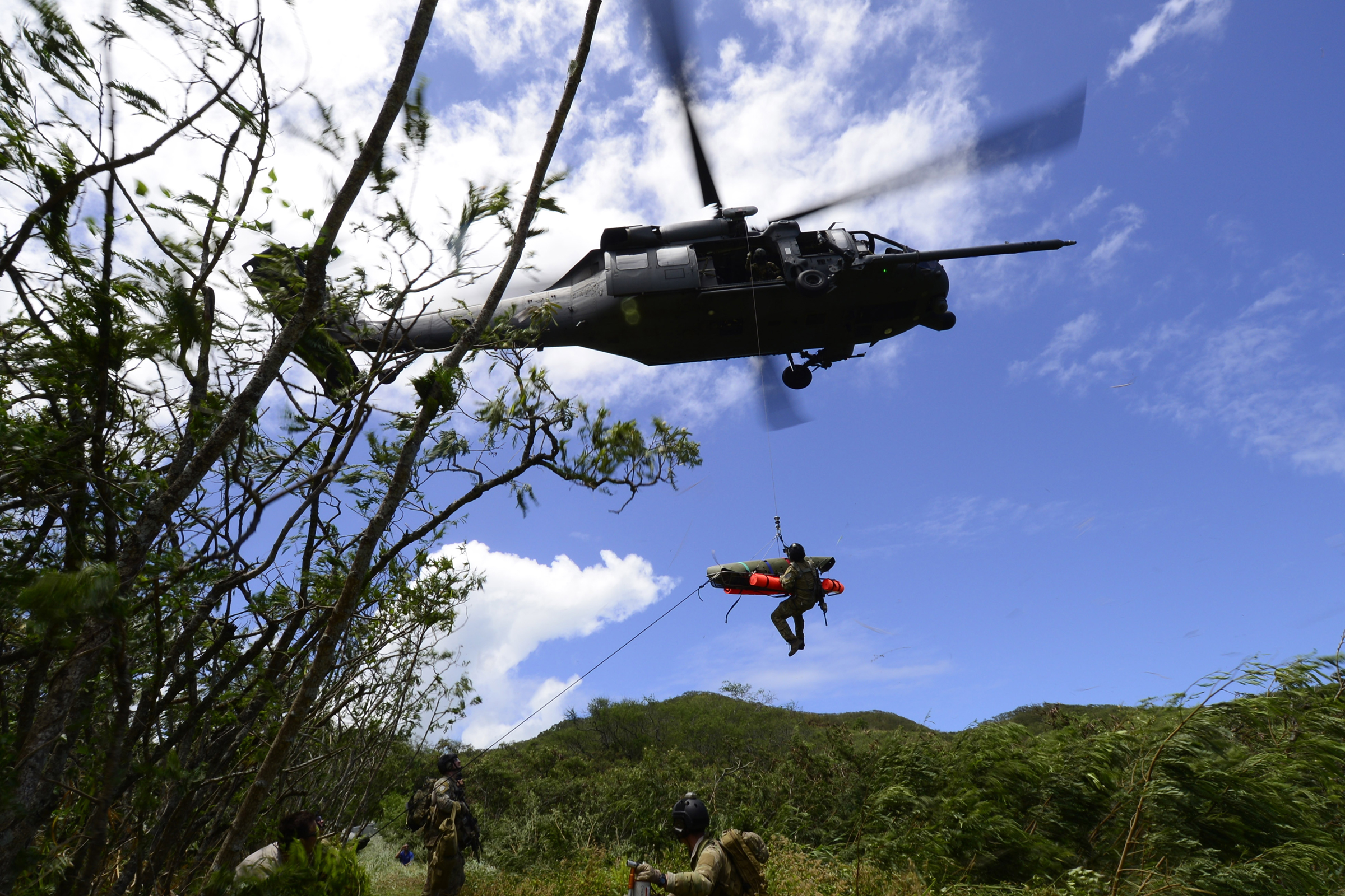 A rescue helicopter hovers above lush, hilly terrain. A man in a helmet descends from a cable with big packs of supplies to join 2 pararescuemen below.